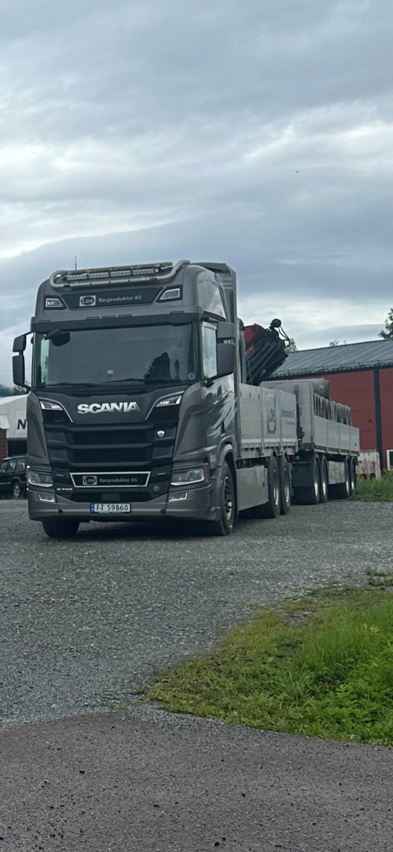 Svart Scania lastebil med flatbed tilhenger parkert på grusplass under skyet himmel, industribygning i bakgrunn